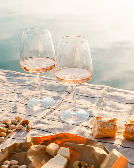 Two glasses of rosé wine beside cheese and nuts on a table on a pier at sunset