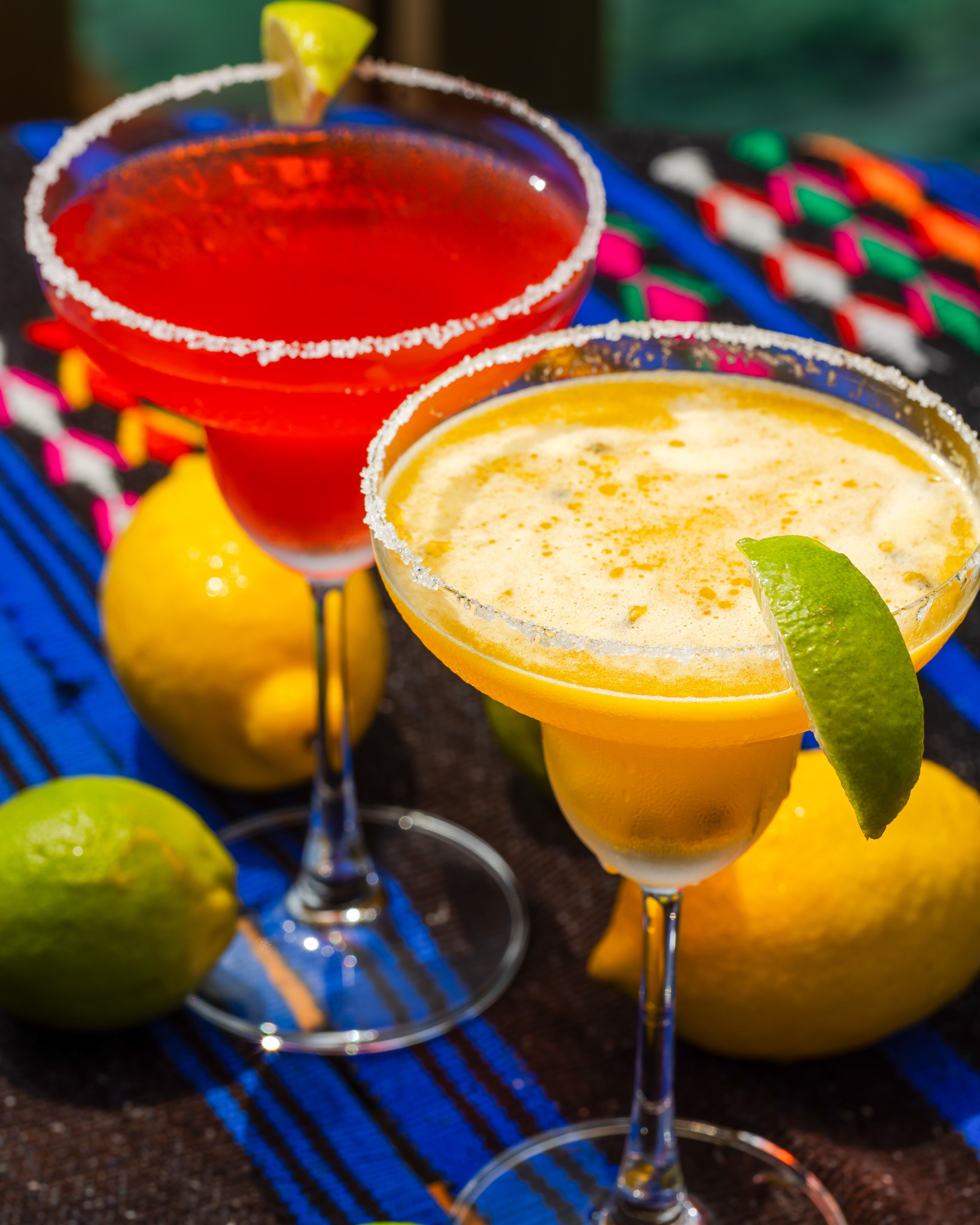 Two margarita cocktails with salted rims, one red and one yellow, beside fresh lemons and limes on a colorful woven cloth