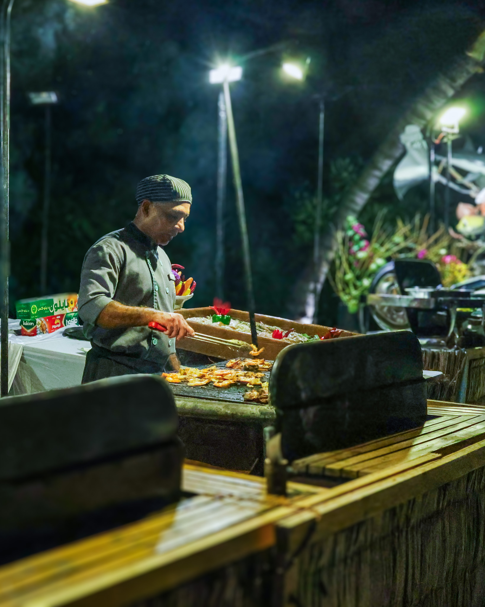 Chef cooking at an outdoor live grill station at night under bright string lights