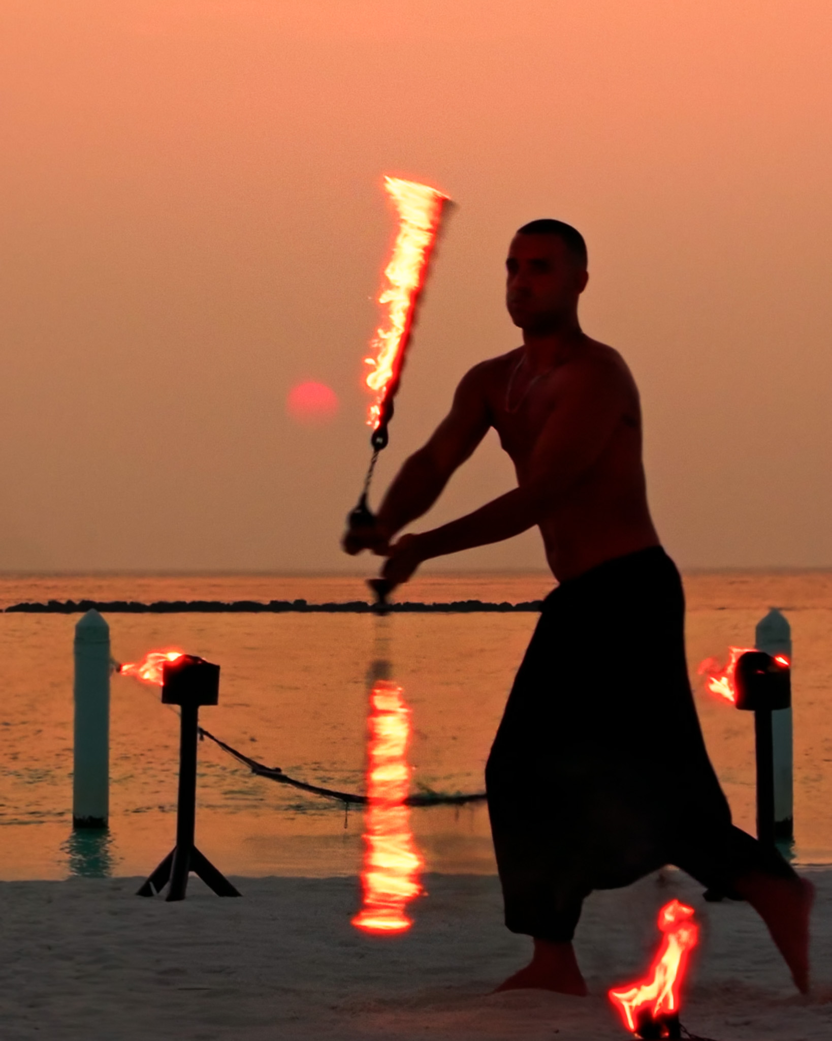Silhouette of a fire performer spinning flaming batons against an orange sunset sky